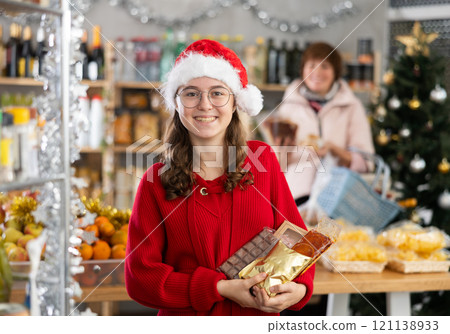 Positive girl in festive hat choosing sweets for New Year's holiday in supermarket with mother choosing food on shelves in 121138933