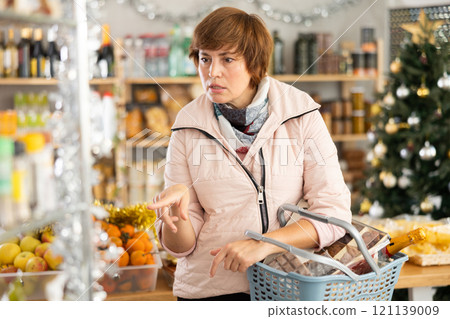 Portrait of attentive female shopper with cart choosing food and wine for Christmas celebration in grocery department of supermarket Portrait of attentive female shopper with cart choosing food and wine for Christmas celebration in grocery department of supermarket 121139009