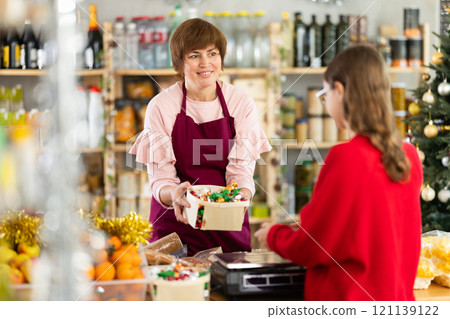 Friendly attentive woman seller in grocery store standing behind counter full of products for Christmas table and weighing and selling candies to buyer girl Friendly attentive woman seller in grocery store standing behind counter full of products for Christmas table and weighing and selling candies to buyer girl 121139122