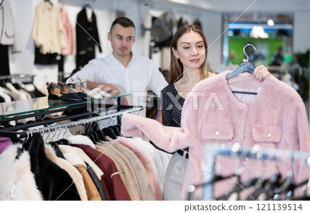 Girl stopped near showcase with outerwear, holds fur coat hanger in hands and examines product Girl stopped near showcase with outerwear, holds fur coat hanger in hands and examines product 121139514