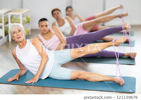 Elderly woman lies on mat and performs stretching exercises with resistance bands in group pilates class Elderly woman lies on mat and performs stretching exercises with resistance bands in group pilates class 121139676