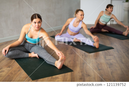 Active fit women in sportswear improving flexibility while practicing Janu Sirsasana stretching position in yoga studio 121139752