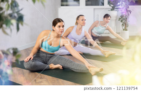 Calm sporty women stretching in Janu Sirsasana and bending head to knee posture during training together in yoga class Calm sporty women stretching in Janu Sirsasana and bending head to knee posture during training together in yoga class 121139754