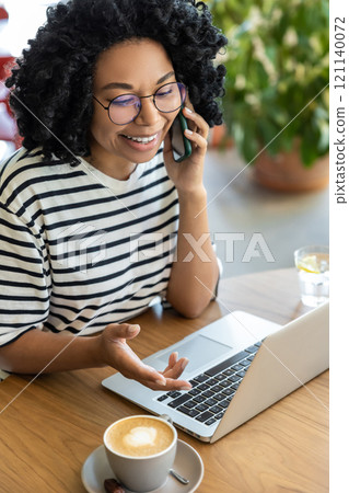 Curly-haired woman working on laptop and having a phone call 121140072