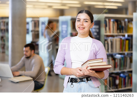 Smiling woman standing with books near bookshelves in library Smiling woman standing with books near bookshelves in library 121140263