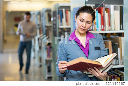 Woman standing alone near bookcase in library browsing textbooks 121140267