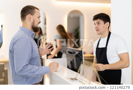 Male stylist stands near barbershop reception desk, communicates with male visitor Male stylist stands near barbershop reception desk, communicates with male visitor 121140279