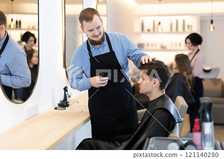 Young man barbershop employee stands next to male client, discussing details of haircut 121140280