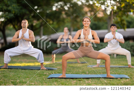 Girl performing Goddess Pose during group yoga in summer park Girl performing Goddess Pose during group yoga in summer park 121140342