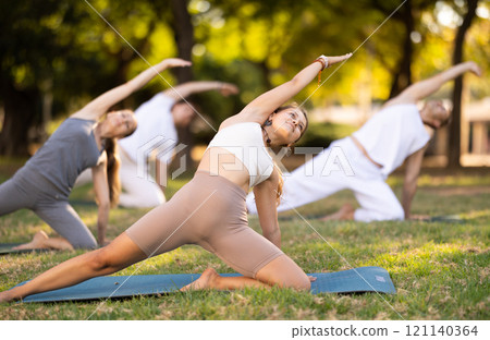 Yoga lesson visitors with girl coach perform Parighasana in park 121140364