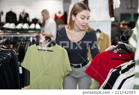 Girl stopped near showcase with outerwear, holds dress hanger in hands and examines product Girl stopped near showcase with outerwear, holds dress hanger in hands and examines product 121142083