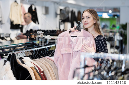 Girl stopped near showcase with outerwear, holds fur coat hanger in hands and examines product 121142118