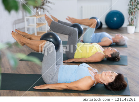 Old woman practicing pilates with softball between legs in light fitness room Old woman practicing pilates with softball between legs in light fitness room 121142261
