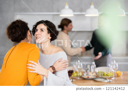 Women hug each other and kiss on the cheek while greeting each other before a festive dinner. Men shaking hands in background 121142412