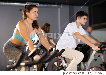 Man and woman taking indoor cycling class at fitness center, doing cardio riding bike Man and woman taking indoor cycling class at fitness center, doing cardio riding bike 121142569