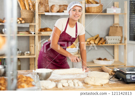Young female baker slicing dough and weighting portioned pieces 121142813