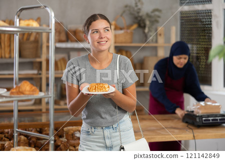 Happy girl buys fresh pastries in an oriental bakery. In background is seller in traditional Muslim clothing 121142849