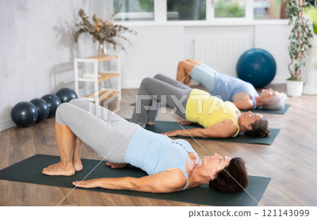 Elderly woman in company of hatha yoga lovers performs bridge pose, setu bandha sarvangasana 121143099