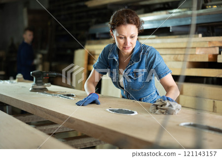 During manufacturing, woman employee grinds surface of wooden board, gives smoothness to surface 121143157