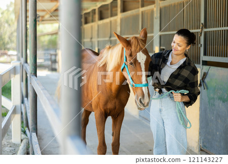 Young woman leading horse out of stable 121143227