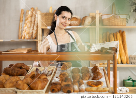 Young woman employee puts croissants in window, arranges display of goods at bakery. Young woman employee puts croissants in window, arranges display of goods at bakery. 121143265