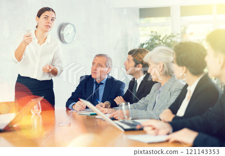 Young woman turning on presentation on projector while office coworkers of different ages making notes at table with laptops Young woman turning on presentation on projector while office coworkers of different ages making notes at table with laptops 121143353