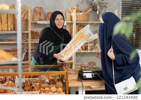 Muslim saleswoman gives baguettes to Muslim female customer Muslim saleswoman gives baguettes to Muslim female customer 121143486