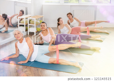 Aged woman exercising with rubber loop band during group pilates Aged woman exercising with rubber loop band during group pilates 121143515