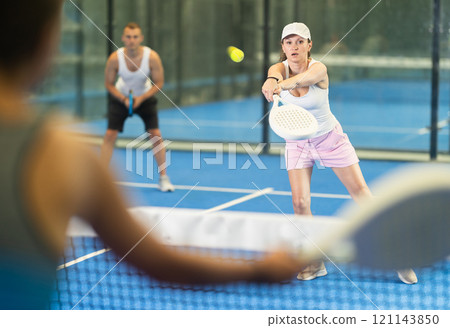 Woman with racket training on indoor court playing padel game and hitting ball 121143850
