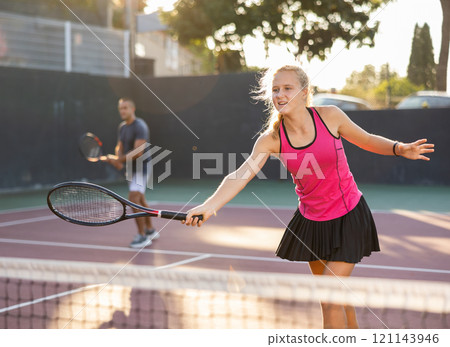 Young woman playing tennis doubles match with male partner on court at sunny day 121143946