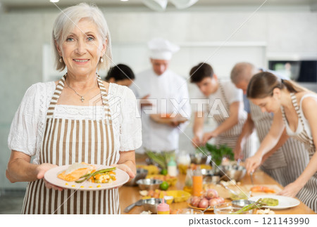 Smiling senior woman holding plate of prepared dish at group cooking class 121143990