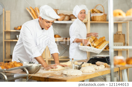 Young man works in bakery as baker, kneads and prepare dough, works with flour. 121144013