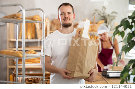 Man buyer with baguettes in package stands near glass showcase in bakery 121144031