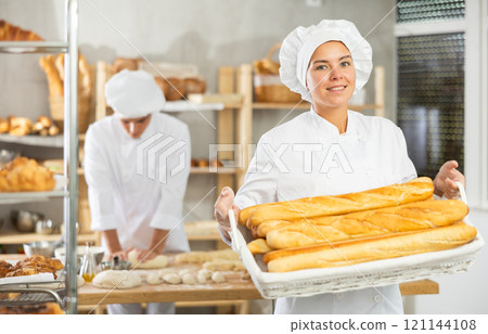 Happy female baker posing with basket of freshly baked baguettes in bakery, demonstrating concept of food industry and baker profession 121144108