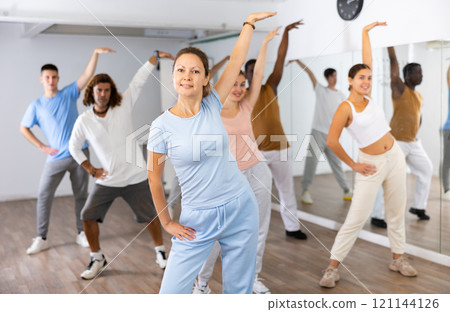 Group of young adult active multiethnic dancers stretching one arm up during learning new dance exercise movement in gym 121144126