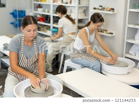 Three young women sits near potter wheel and makes clay crafts 121144253