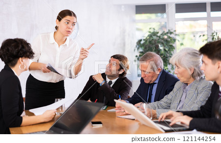 Smart young woman sharing ideas in front of office colleagues of different ages taking notes at table with laptops 121144254