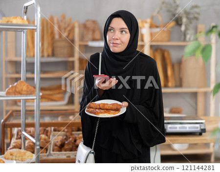 European Muslim girl customer holds pastry on disposable plate and recognizes it using app 121144281