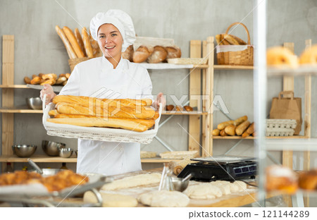 Smiling female baker presenting fresh baguettes in bakery 121144289