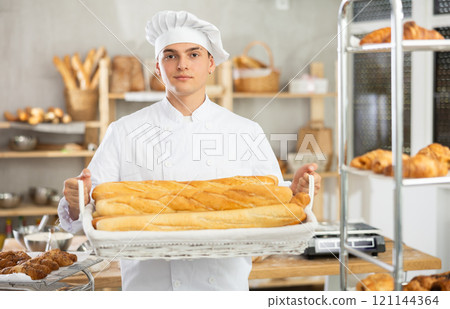 Young male baker holding baguettes in wicker basket Young male baker holding baguettes in wicker basket 121144364