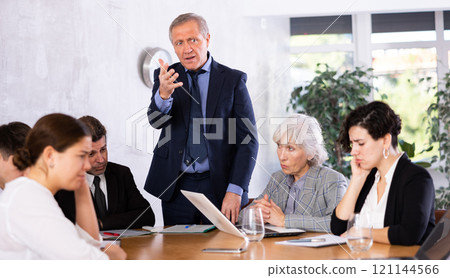 Angry elderly male boss scolding office workers of different ages using notebooks at table in office 121144566