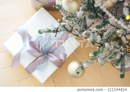 Festively wrapped gift box with bow under beautifully decorated Christmas tree on wooden floor Festively wrapped gift box with bow under beautifully decorated Christmas tree on wooden floor 121144824