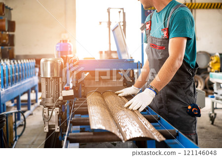 A factory worker is making a metal profile for a roof. Worker at factory measures metal profiled sheet. He works in metal production industrial. metal roofing production, production of roof elements A factory worker is making a metal profile for a roof. Worker at factory measures metal profiled sheet. He works in metal production industrial. metal roofing production, production of roof elements 121146048
