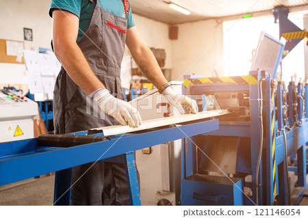 A factory worker is making a metal profile for a roof. Worker at factory measures metal profiled sheet. He works in metal production industrial. metal roofing production, production of roof elements 121146054