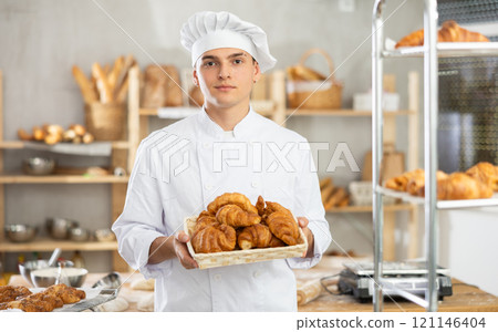 Young male baker holding croissants in square wicker basket Young male baker holding croissants in square wicker basket 121146404