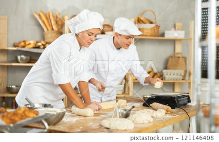 Girl and guy bakers cut small pieces of dough from large lump and put them on scales. 121146408