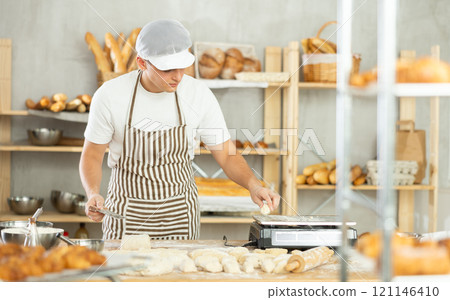 Young male baker cutting and weighing dough on scales in bakehouse 121146410