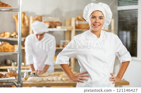 Young baker girl is standing near work table in kitchen, working with dough 121146427