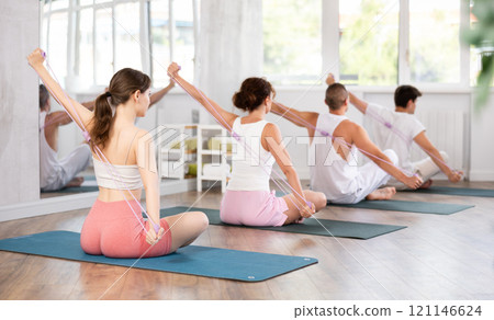Back view of young girl with group of young adults maintaining physical health practicing pilates with resistance band sitting on gymnastics mat at fitness center 121146624