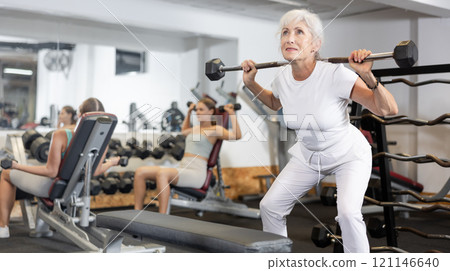 Senior woman trains muscles of arms and back using barbell and differentiated weighting in gym 121146640